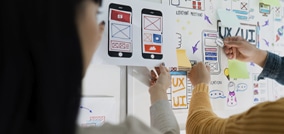 Dois designers trabalhando em um quadro de ideias com esboços de interfaces móveis e conceitos de UX/UI