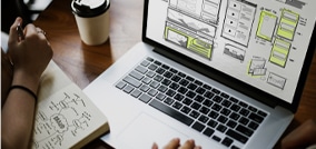 Designer trabalhando no planejamento de uma interface em seu laptop, com esboços de wireframes na tela e anotações em um caderno
